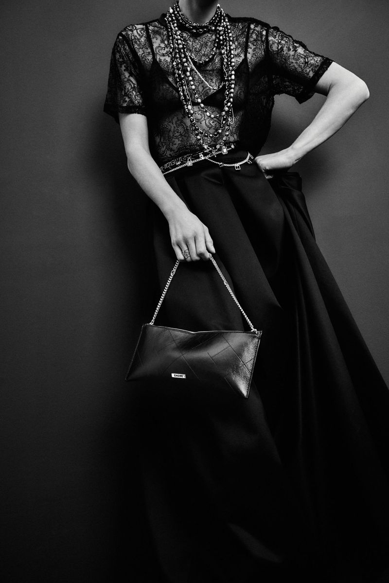 Black and white fashion portrait of a female model wearing a lace top, beads and a long skirt, holding a small black handbag.