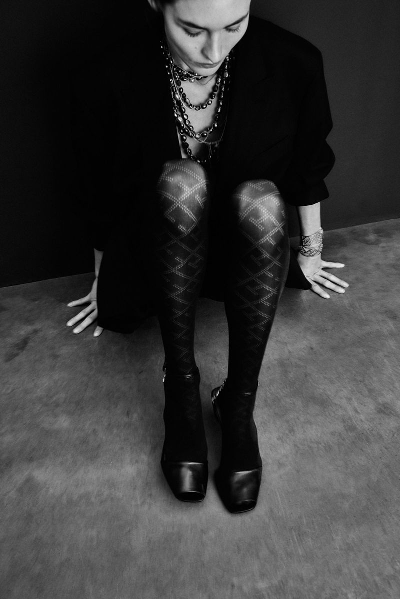 A woman wearing patterned stockings, a jacket, boots, and multiple necklaces poses indoors in a black and white fashion shot.