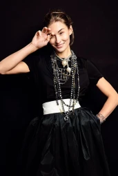 Grace Elizabeth wearing a black t-shirt, white belt, and layered pearl necklaces against a dark background