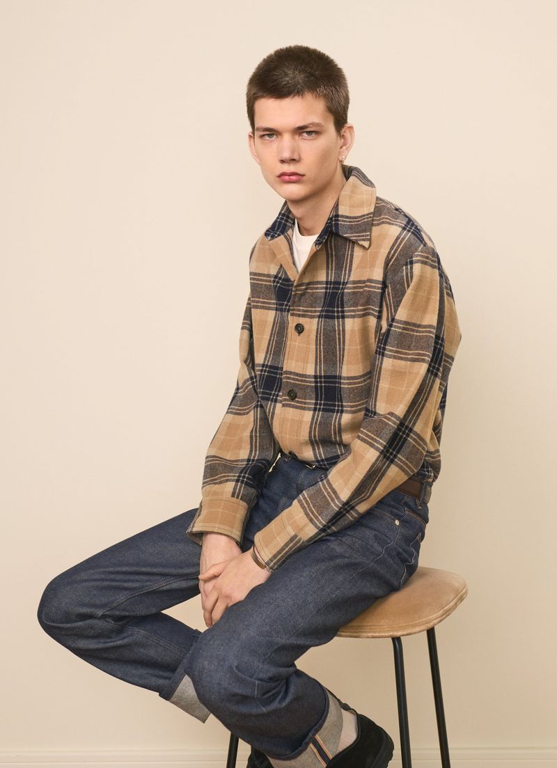 Male model wearing a plaid jacket and jeans, sitting on a stool in a studio setting. Neutral background, natural lighting, fashion editorial.