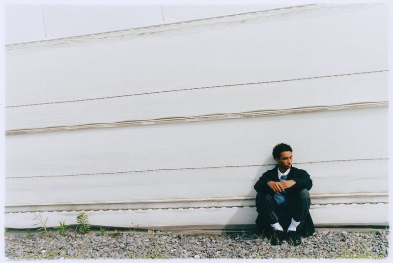 Man in business attire sits against a white corrugated metal wall, in a portrait-style composition with natural light and outdoor setting.