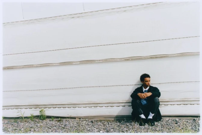 Man in business attire sits against a white corrugated metal wall, in a portrait-style composition with natural light and outdoor setting.