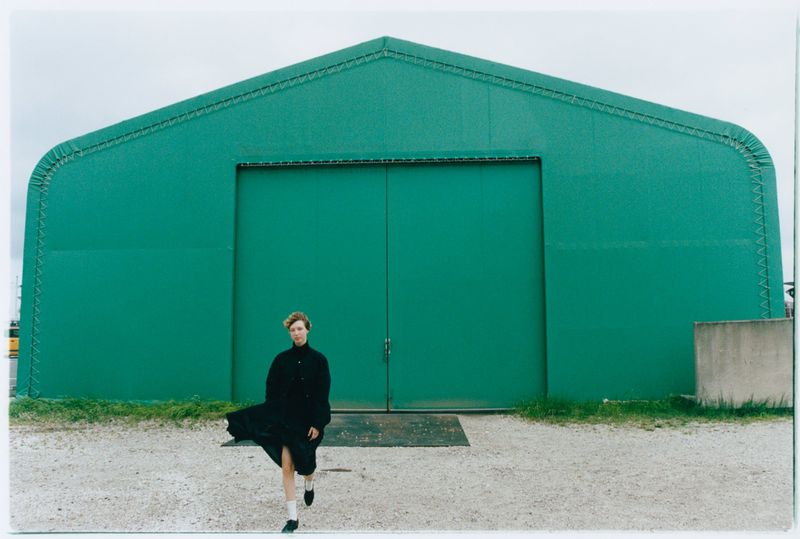 Woman in black coat walks confidently in front of a green warehouse on a cloudy day. A fashion-editorial outdoor photograph.