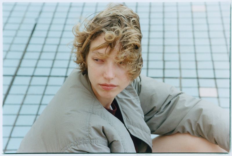 Close-up portrait of a young adult woman with curly blonde hair wearing a jacket against a tiled grid background. Fashionable and modern.