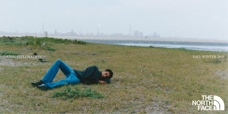 Adult male model wearing jeans and a dark jacket lies on a grassy field near the water and an industrial cityscape in the background.