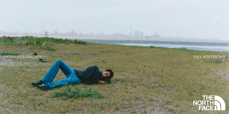 Adult male model wearing jeans and a dark jacket lies on a grassy field near the water and an industrial cityscape in the background.