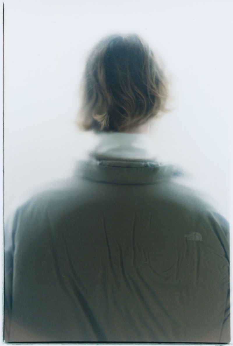 Person with wavy hair and a turtleneck wearing a beige puffy jacket facing away in a bright white photo studio setting.
