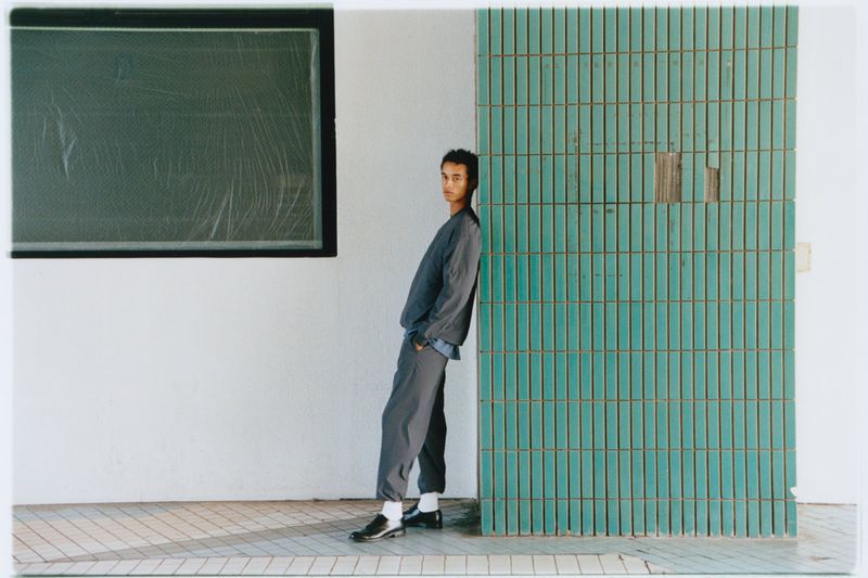 Adult male in casual clothing leans against a tiled wall next to a dark-framed window in an indoor, urban environment in a medium shot.