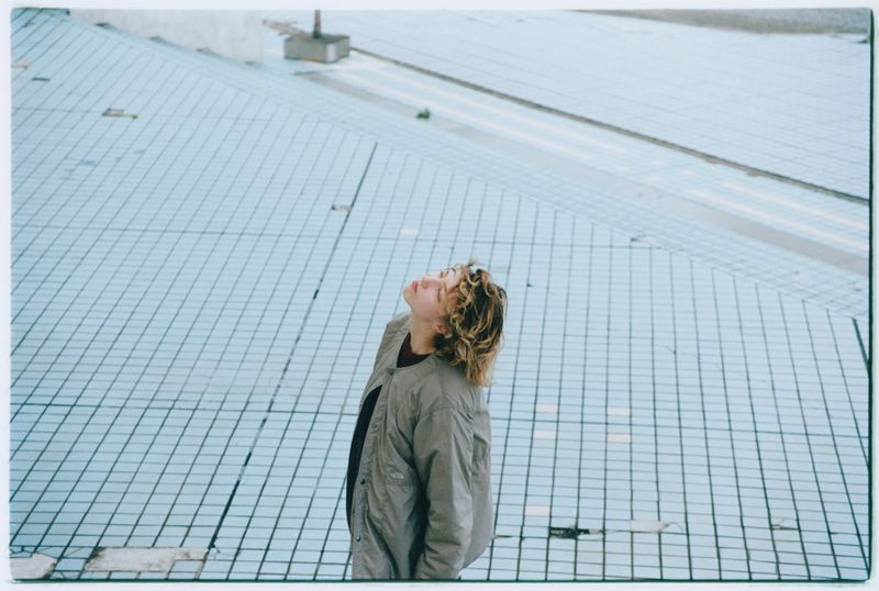 Young adult gazes upward on a grid surface. Overhead shot reveals tan jacket, curly hair against pale grid, emphasizing depth.
