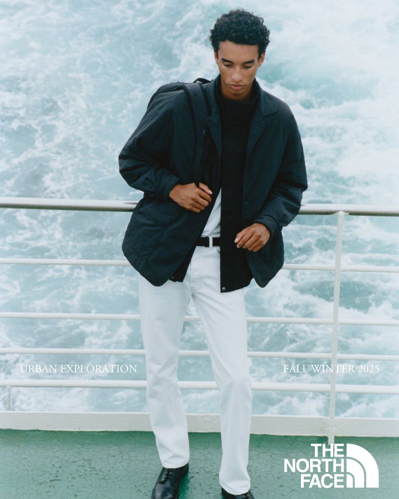 Fashion shot featuring a young adult male in an urban outdoor setting, wearing a black jacket, white pants, and black leather shoes.