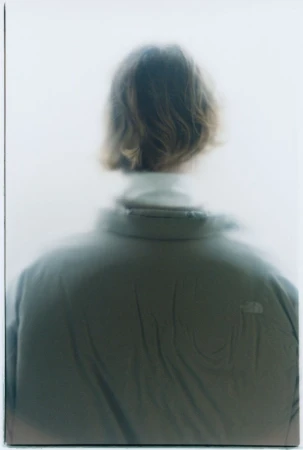 Back view of a person with wavy hair wearing a grey high-collar jacket against a soft, pale, misty background.