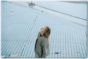 A person with curly blonde hair wearing a grey jacket stands on a pale blue tiled surface looking up.