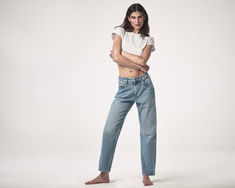 Full-length studio shot of a young adult woman with arms crossed, wearing a white t-shirt and light blue denim jeans on a white background.