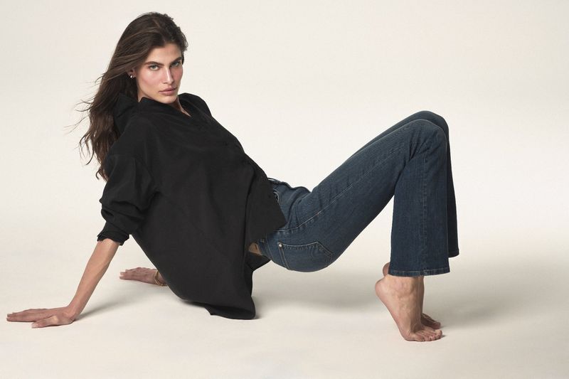 Woman in blue jeans and a black shirt lying down in a studio setting with a soft white background in a fashion pose.