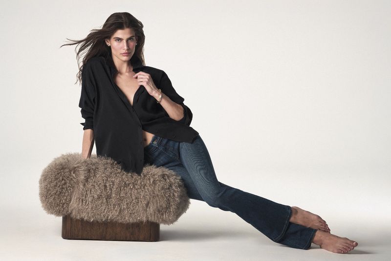 A brunette woman with long hair models black shirt and blue jeans, seated on a furry stool in front of a neutral backdrop.