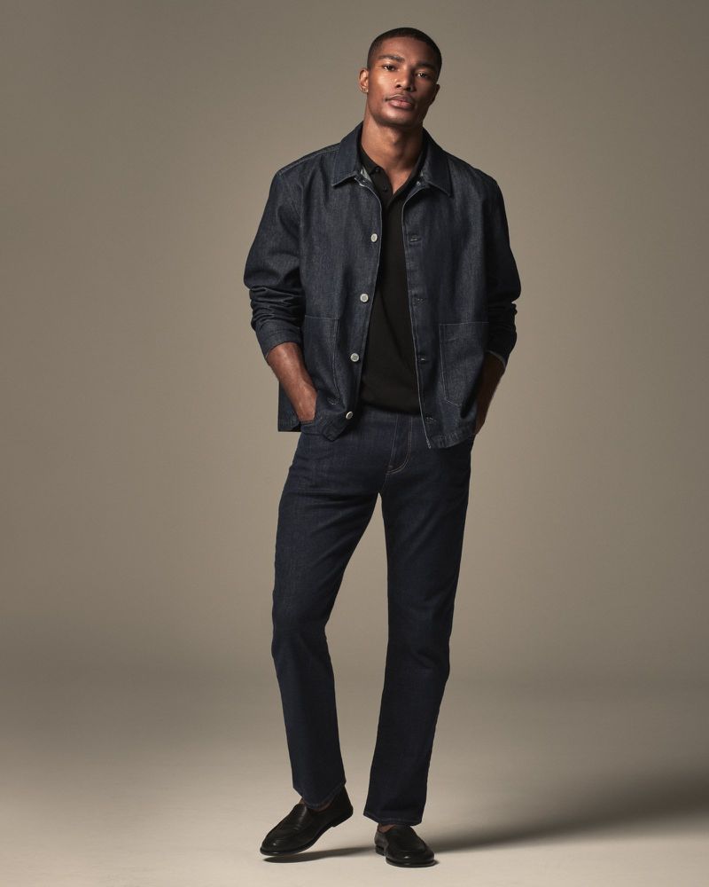 Full body shot of an adult Black male in a denim jacket, black polo shirt, jeans, and dress shoes, posing in a studio setting.