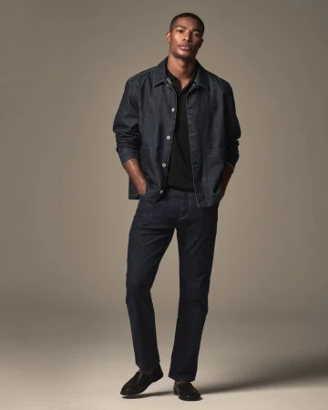 Full body shot of an adult Black male in a denim jacket, black polo shirt, jeans, and dress shoes, posing in a studio setting.