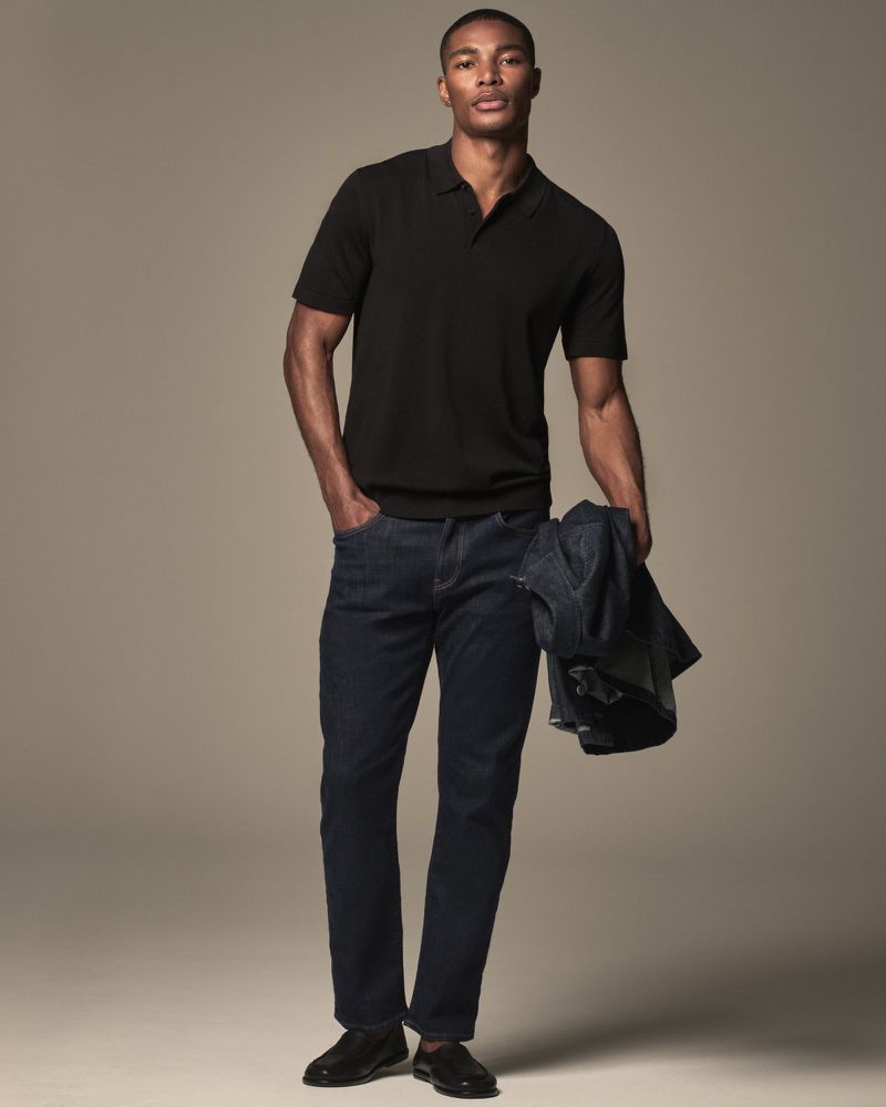 Full-length shot of an African American man in jeans and a black polo shirt, holding a jacket in studio setting. Fashion editorial.