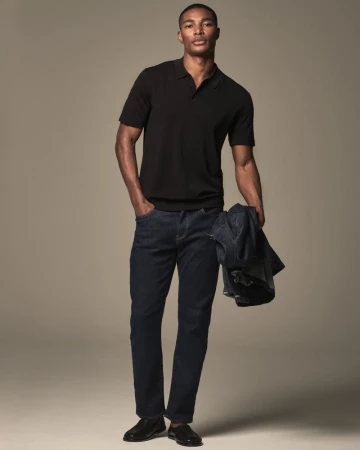 Full-length shot of an African American man in jeans and a black polo shirt, holding a jacket in studio setting. Fashion editorial.