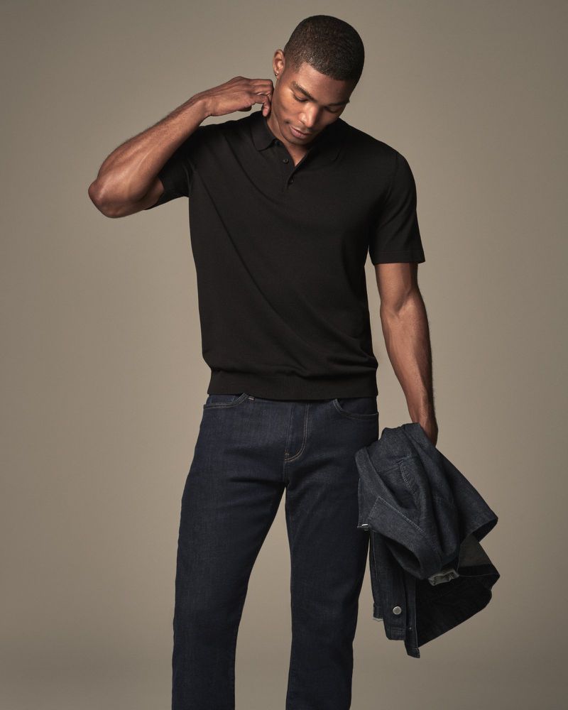 A stylish dark-skinned man in a black polo shirt and jeans poses in a studio with a denim jacket in hand, looking down with a neutral expression.