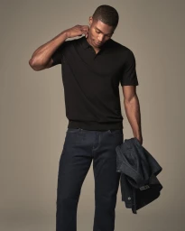 A man wearing a black short-sleeved polo shirt and dark indigo jeans holding a denim jacket against a neutral studio backdrop.