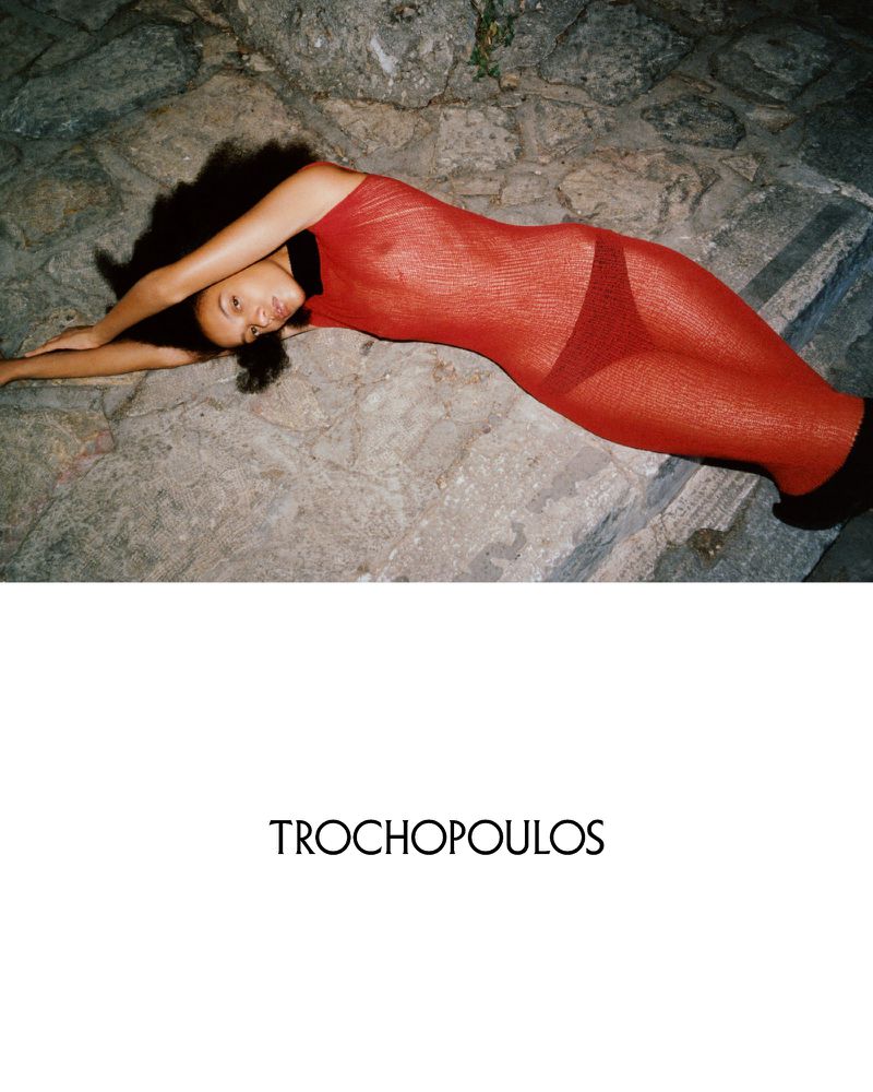 Woman in red sheer dress lying on stone surface. Fashion shot with shadows, textured background, and visible designer logo.
