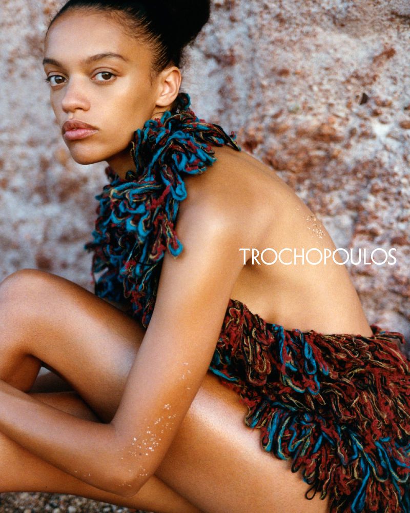 Adult woman in a fringed dress poses against a rock face, the shot is brightly lit and shows her from the waist up.