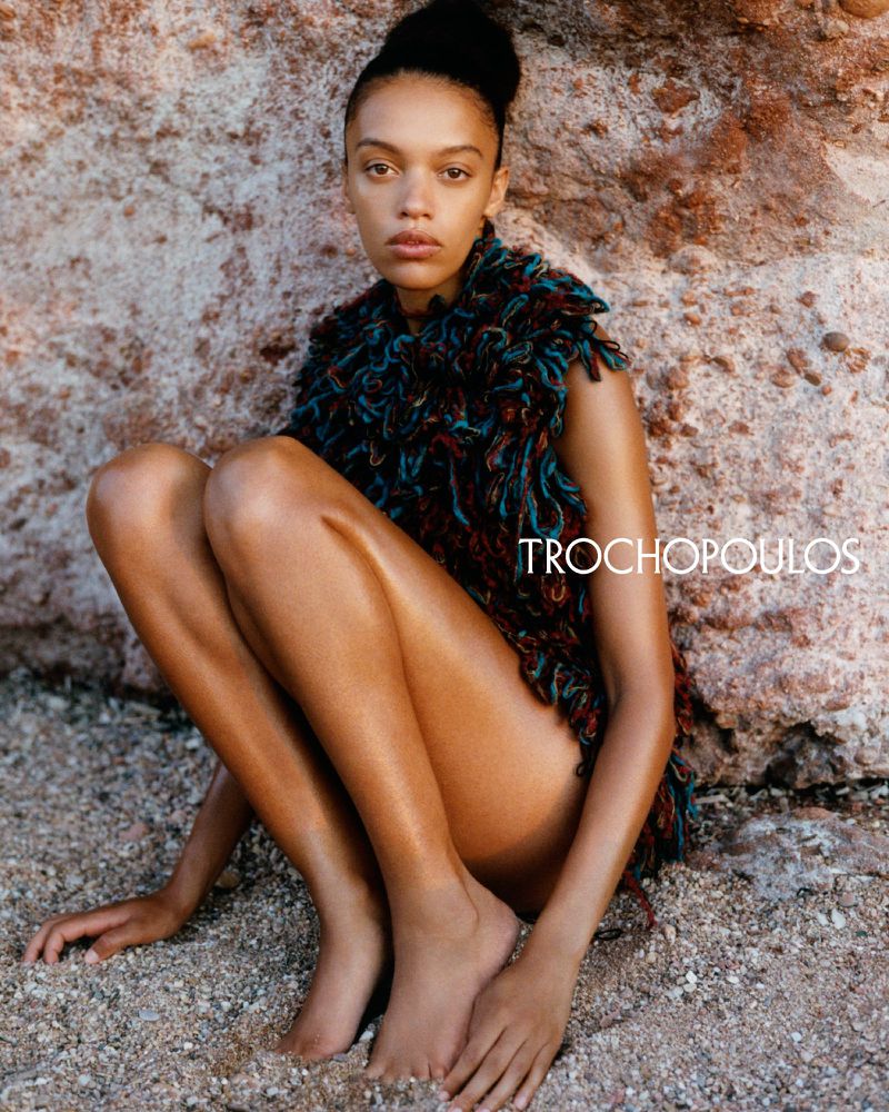Young woman with dark hair in a textured top crouches on a sandy surface, backed by a textured rock wall, creating a natural fashion portrait.