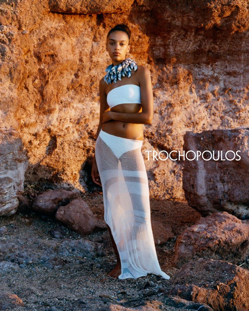 A woman in a white bikini and a sheer cover-up stands on a rocky beach with a textured rock formation behind her in natural light.
