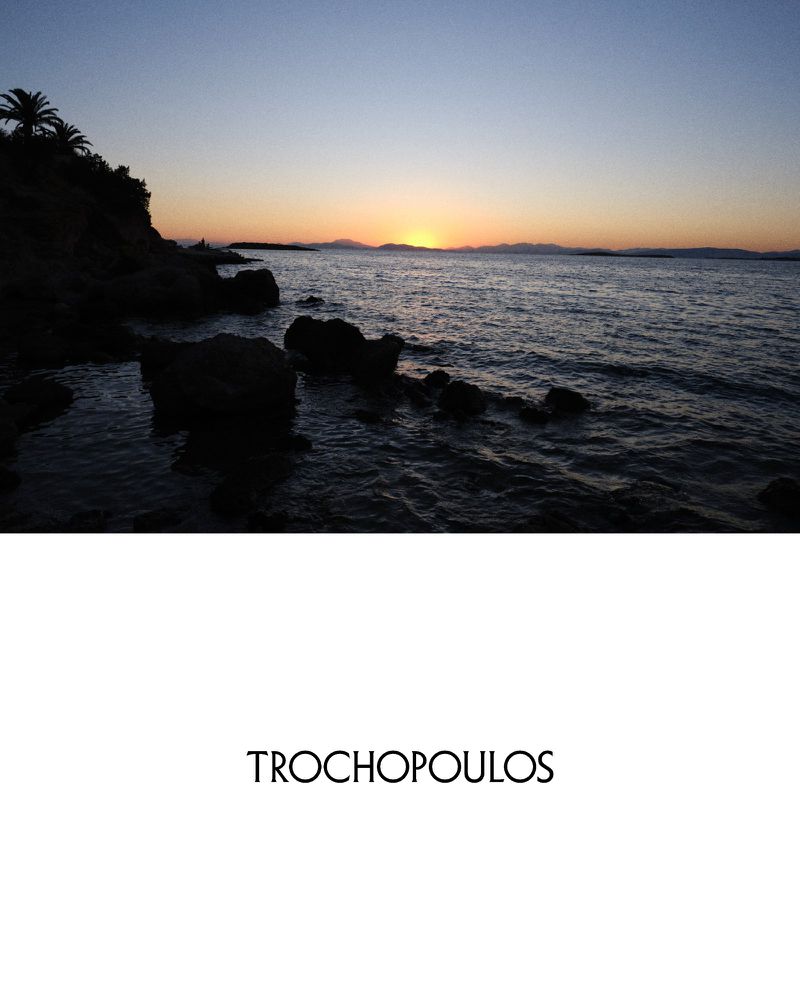 Coastal sunset with dark rocks and gentle waves, silhouetted trees on the cliff, and soft light illuminating the sky.