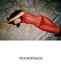 Model lying on rough stone ground wearing a translucent red dress with black footwear for Trochopoulos F/W 2025 campaign.