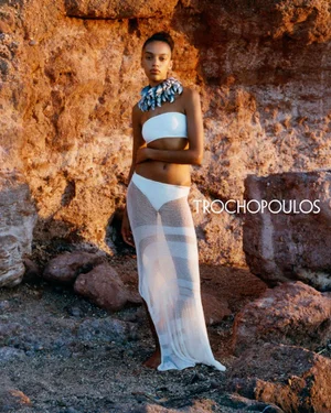 Model in white swimwear and sheer skirt posing against rocky terrain for Trochopoulos F/W 2025 campaign.