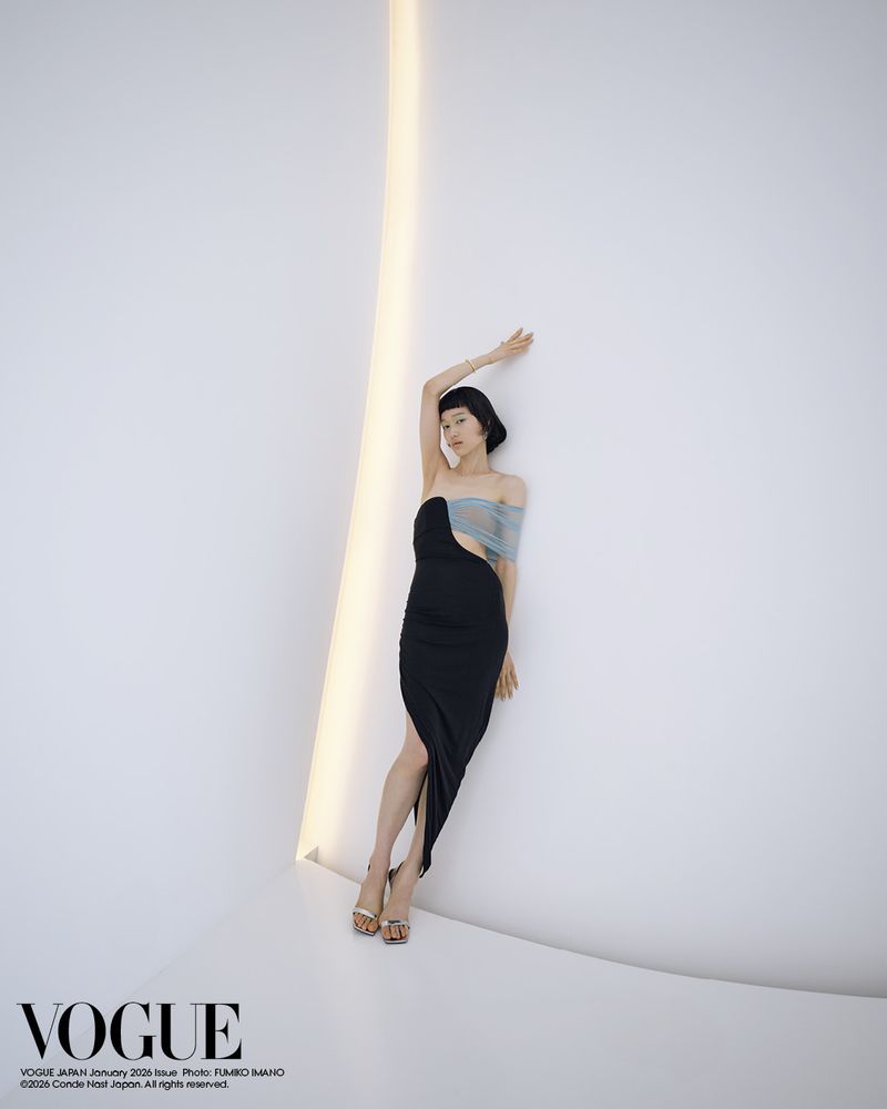 An Asian woman poses in a black dress with sheer blue fabric, leaning against a curved white wall with a light beaming from above.