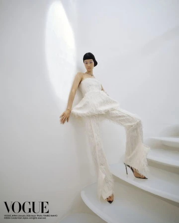Fashion editorial shot: a slender Asian woman posing in a white fringed strapless top and pants on a white staircase indoors.