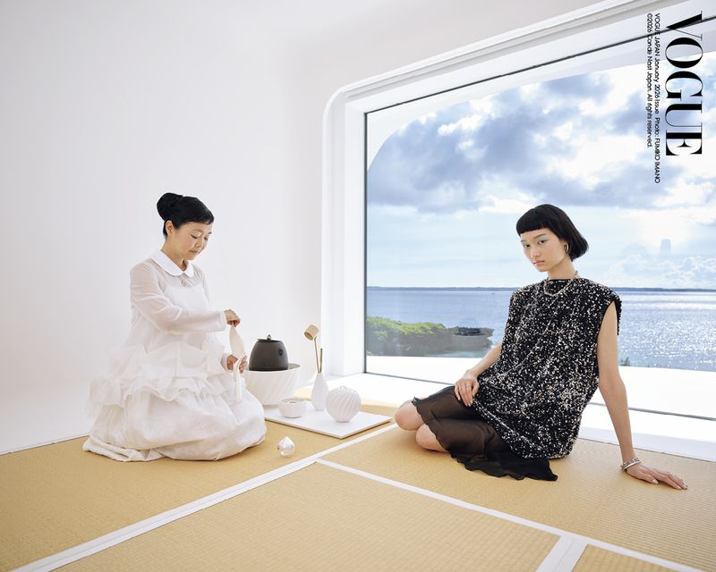 Two women in elegant dresses share a serene space overlooking a sea view from a large window, creating a minimalist fashion editorial.
