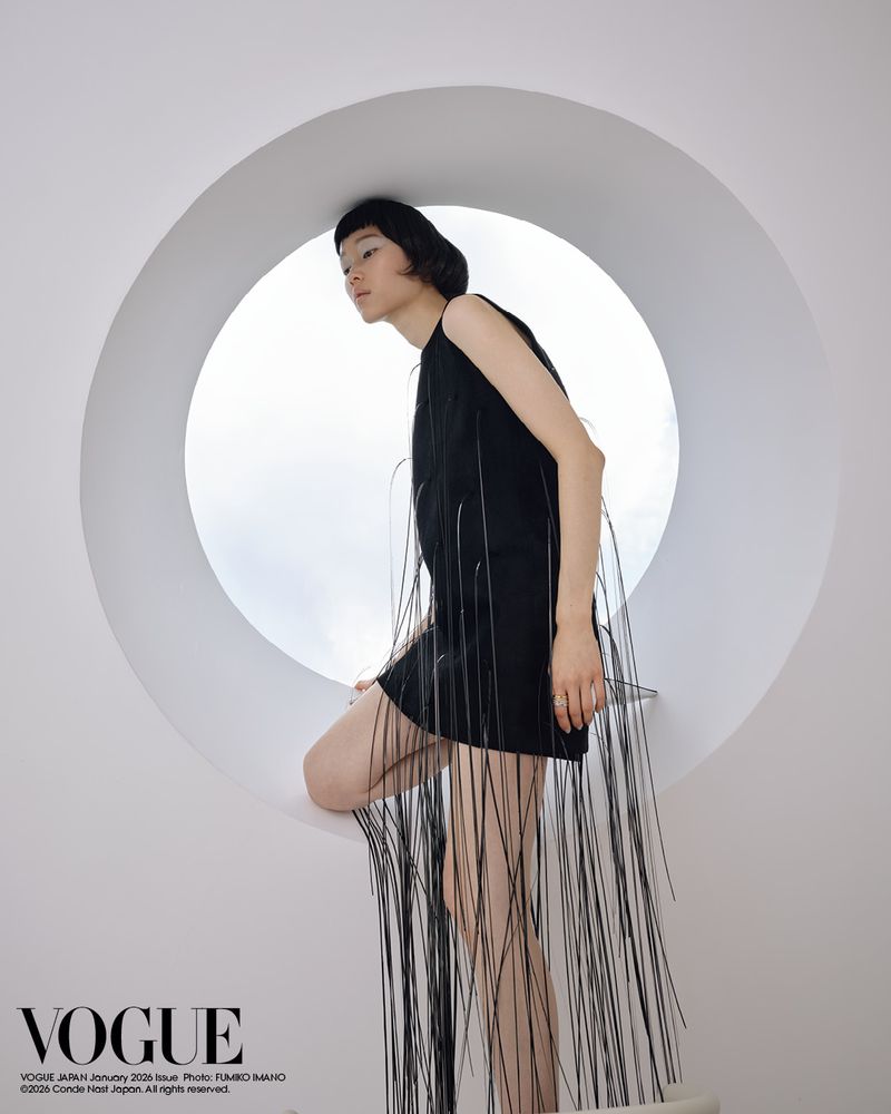A model in a black dress poses in front of a circular opening in a bright white architectural space, showcased in a fashion editorial.