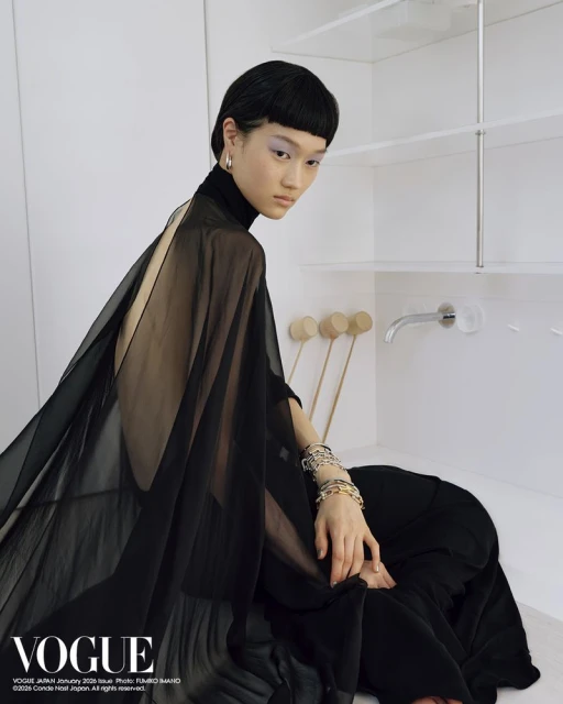 Asian woman in a black dress posing in front of white shelves and a bathroom sink with a vogue magazine logo in the corner.