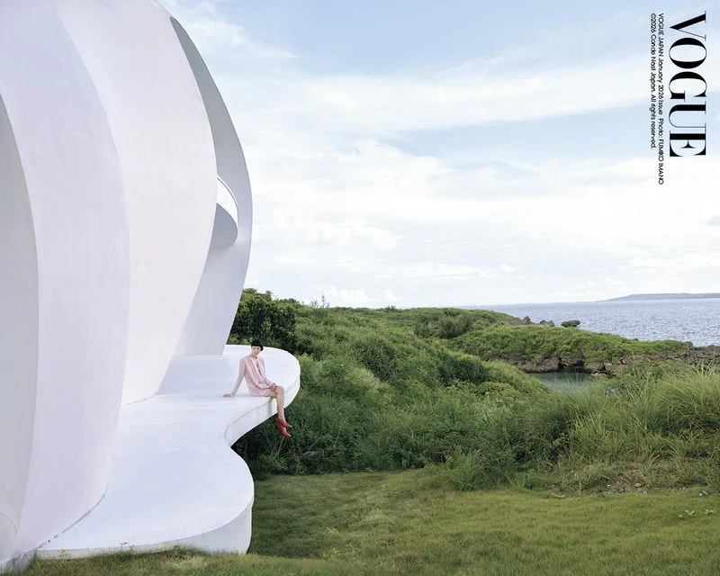 Adult woman in a pink dress sits on the edge of a white building with green foliage and a body of water in the distant background.