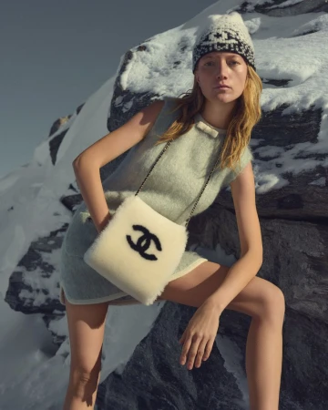 A female model poses in a sleeveless dress and Chanel bag with snow-covered rocks in the background for a fashion editorial.