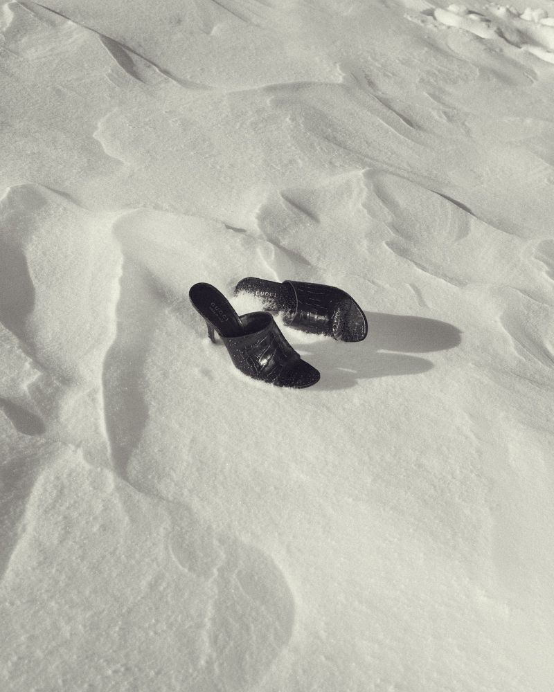 Black heeled mules rest on textured white snow, creating a monochrome fashion still life with an emphasis on contrasting textures.