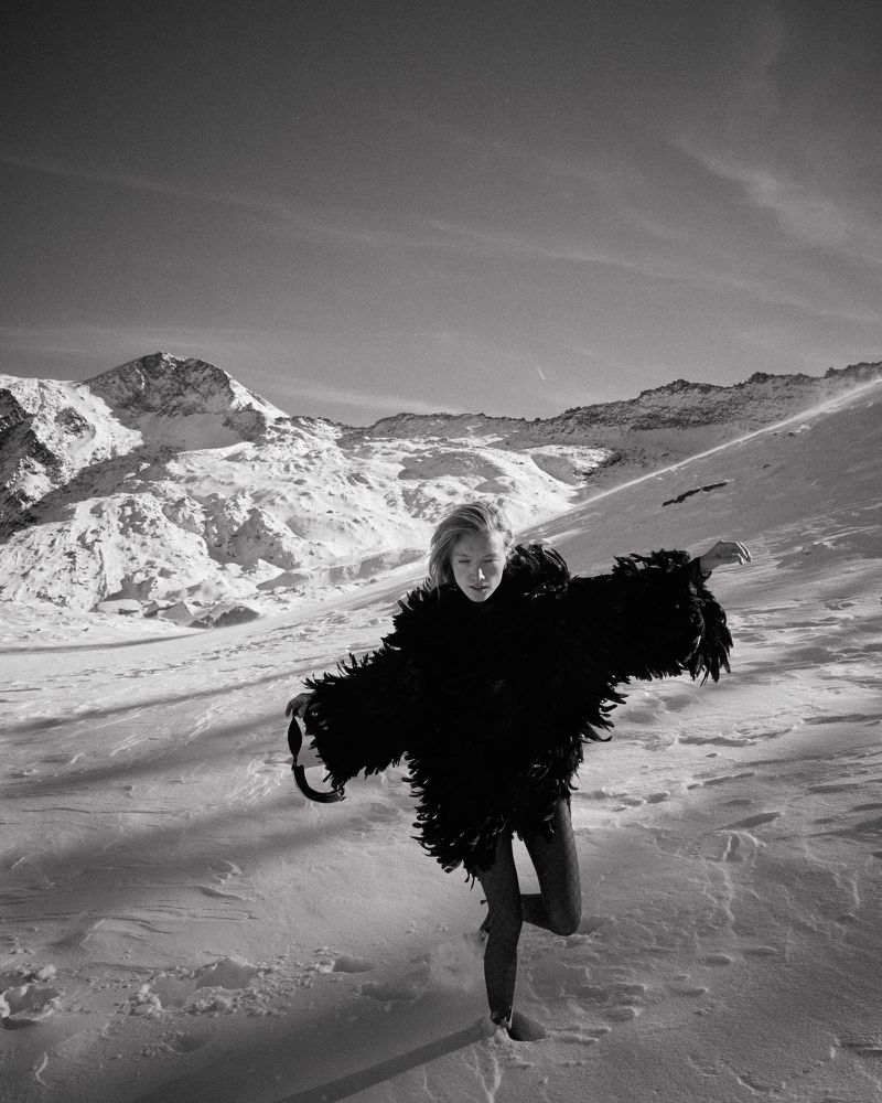 A fair-skinned woman in a feather coat poses in a snowy, mountainous landscape on one leg. Shot is monochrome, emphasizing textures and contrasts.