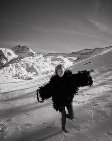 A fair-skinned woman in a feather coat poses in a snowy, mountainous landscape on one leg. Shot is monochrome, emphasizing textures and contrasts.