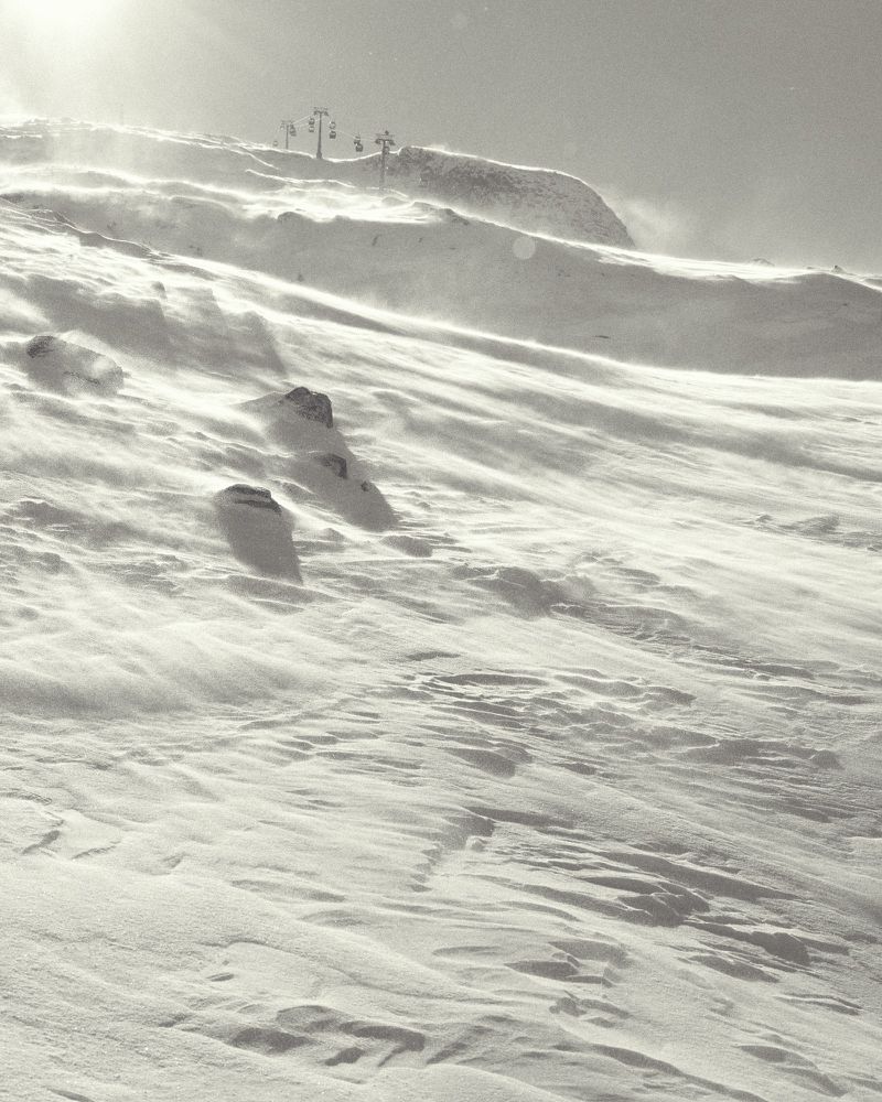 Snowy mountain landscape with wind blowing snow and a ski lift in the background, monochromatic scenery.
