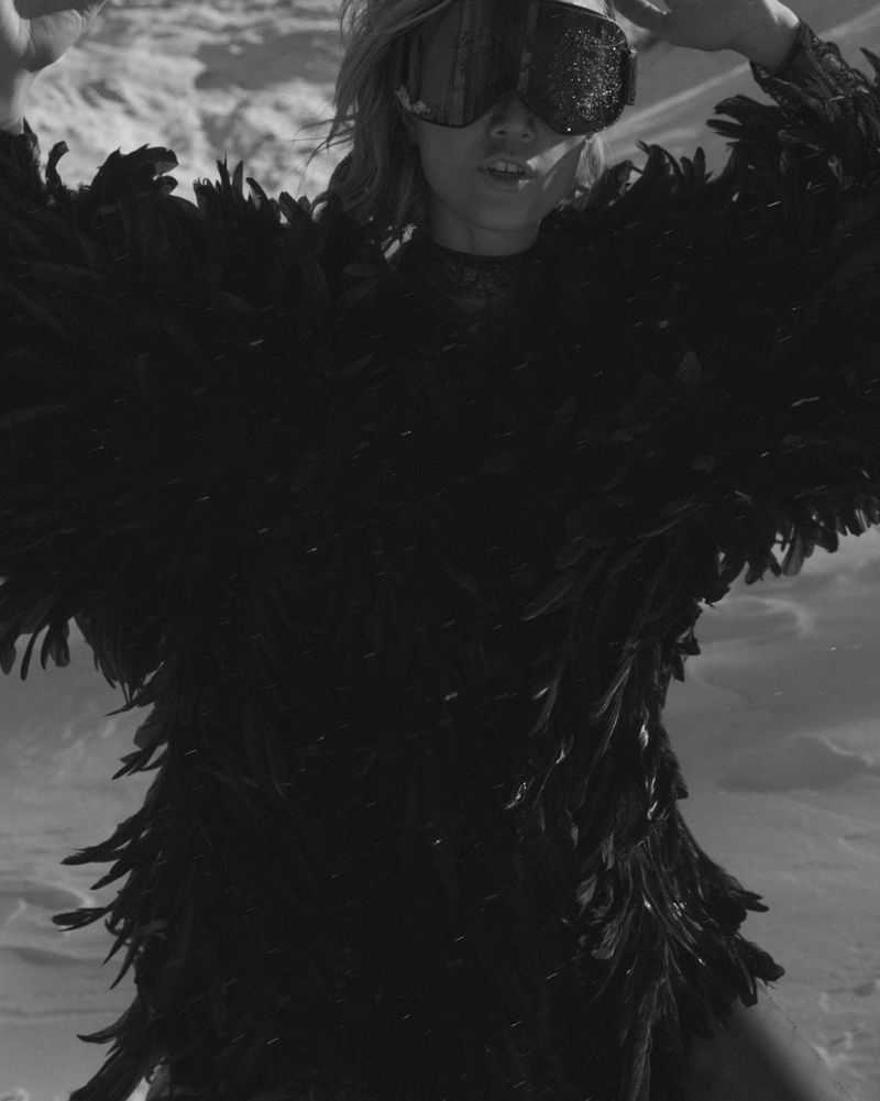 Black and white medium shot of a woman wearing ski goggles and a black feathered garment in a snowy landscape.