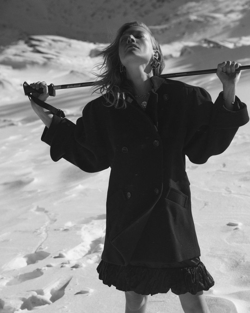 Woman poses in black jacket and dress against snowy mountain backdrop, holding ski poles overhead in a black and white fashion shot.