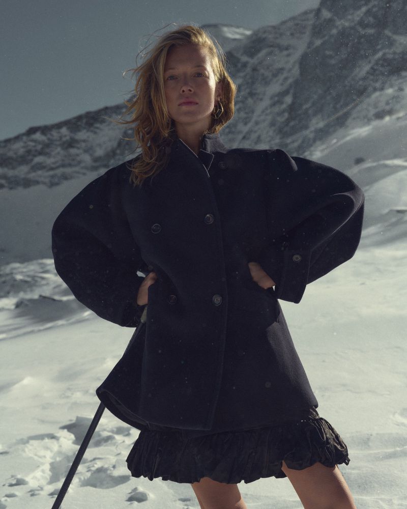 A blonde woman in a black coat and skirt poses in a snowy mountain landscape, standing with hands on hips and mountains behind her.