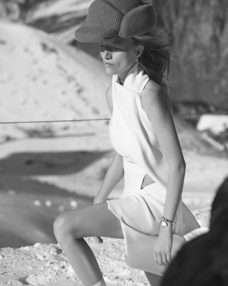 Black-and-white fashion shot of an adult woman wearing a white dress and hat, posing against a snowy, mountainous backdrop on a sunny day.