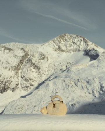 Luxury handbag displayed in a snowy mountain landscape with clear blue sky, showcasing fashion and nature with neutral color palette.