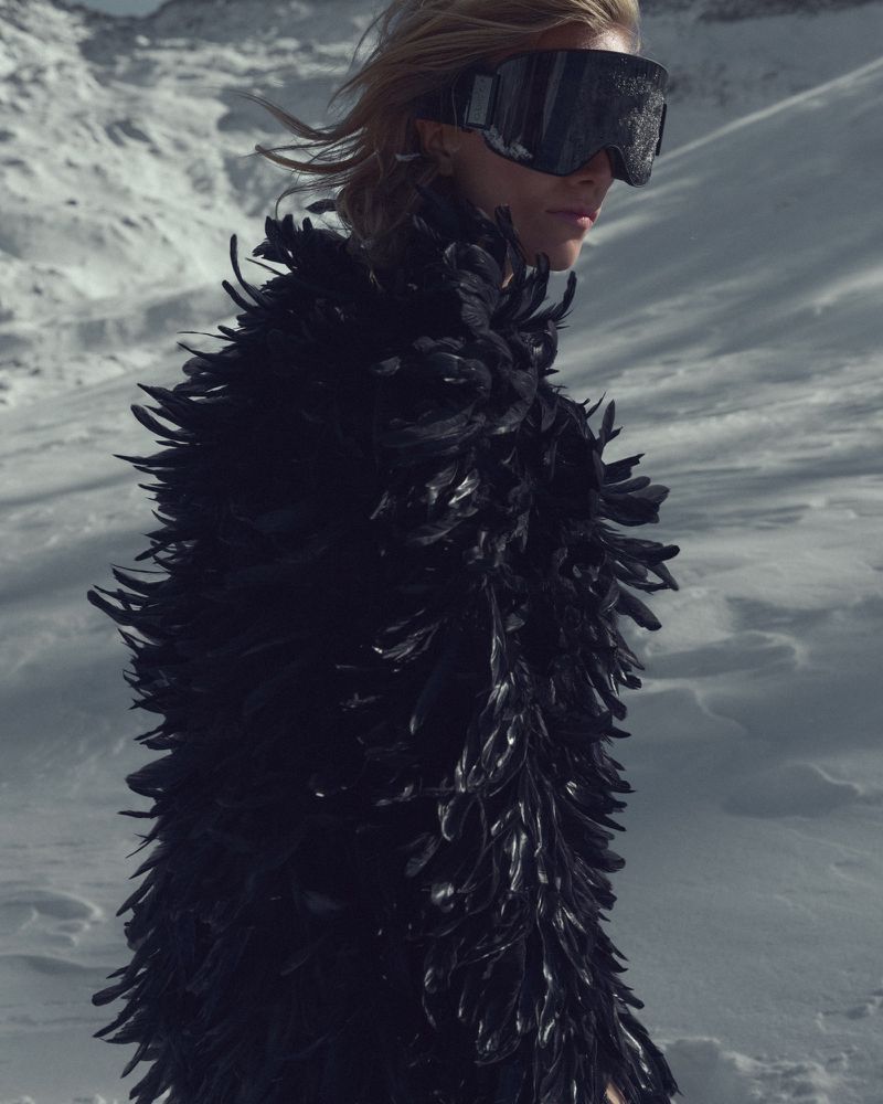 Fashion shot of a blond woman wearing large ski goggles and a black feather coat in a snowy mountain landscape on a sunny day.