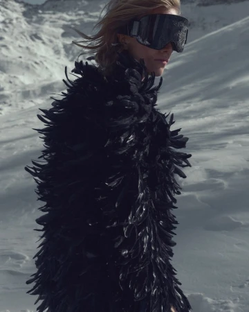 Fashion shot of a blond woman wearing large ski goggles and a black feather coat in a snowy mountain landscape on a sunny day.